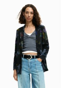 Blazer nero con motivo floreale, top grigio, jeans a gamba larga azzurri e cintura nera con fibbia geometrica argento.