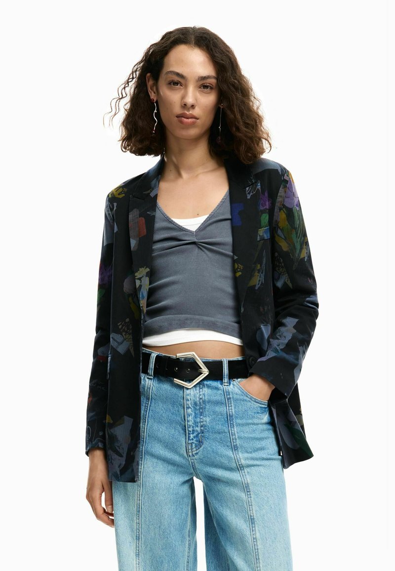 Blazer nero con motivo floreale, top grigio, jeans a gamba larga azzurri e cintura nera con fibbia geometrica argento.