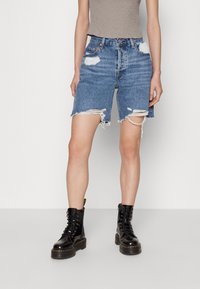 Jeansshorts i medelblått, med slitna detaljer och fransad nederkant. Matchas med en åtsittande grå linne och svarta snörboots.