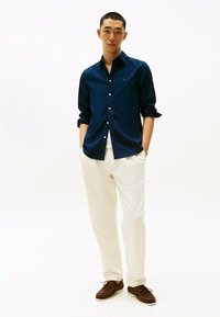 Chemise bleu marine à boutonner, manches retroussées, associée à un pantalon crème et des mocassins marron foncé. Design simple, texture lisse, branding minimal.
