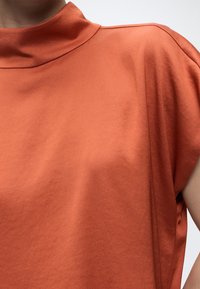 Haut orange à manches courtes avec un col montant, porté par une personne visible du cou jusqu'au milieu du torse sur un fond uni.