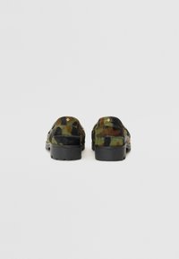 Et par camo-mønstret slip-on sko med sorte gummisåler, vist fra baghøjden på en ensfarvet hvid baggrund.