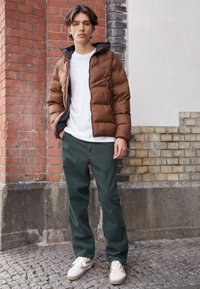 Junger Mann, der eine braune Steppjacke, ein weißes T-Shirt, dunkelgrüne Hose und weiße Turnschuhe trägt und vor einer Wand aus Ziegeln und Fliesen steht.