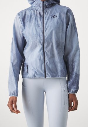 Person trägt eine hellblaue Kapuzenjacke mit Reißverschluss und passende hellblaue Leggings mit einer Reißverschlusstasche an der Seite mit der Aufschrift "Nike Trail."