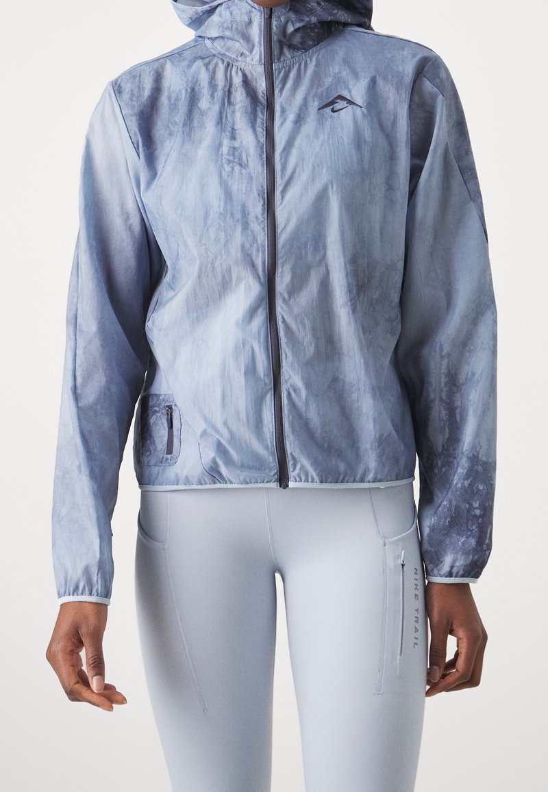 Personne portant une veste à capuche bleu clair zippée et des leggings bleu clair assortis avec une poche latérale zippée étiquetée "Nike Trail."