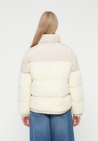 Pufferjack met een hoge kraag, crème schapenvacht textuur aan de onderkant, en een gladde beige bovenkant, met elastische manchetten.