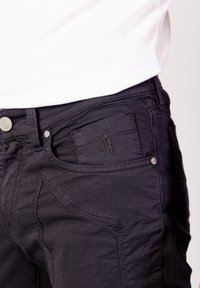 Vaqueros de mezclilla negra con un bolsillo delantero con logo bordado, herrajes plateados y costuras distintivas en los laterales. Diseño de ajuste casual.