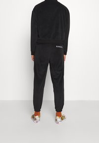 Chándal de velour negro con puños y cintura acanalados, con el logo 'KANGOL' en la parte trasera de la chaqueta y los pantalones. Modelo lleva zapatos estampados.