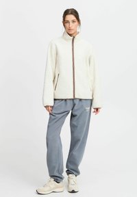 Giacca in pile color crema con colletto alto e motivo a diamante, abbinata a pantaloni felpati grigi larghi e sneakers beige. Chiusura con zip e tasche laterali.