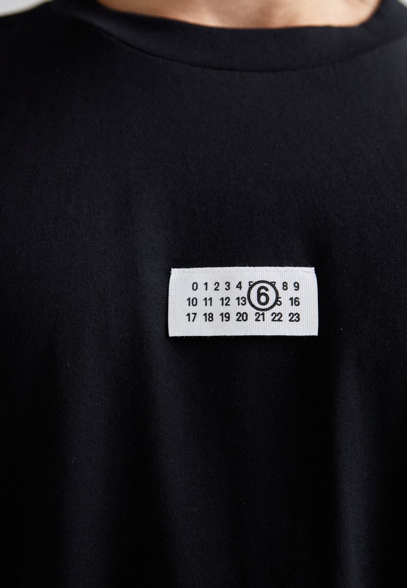T-shirt in cotone nero con un'etichetta bianca con numeri e un design circolare. Il tessuto appare morbido, con una texture liscia.