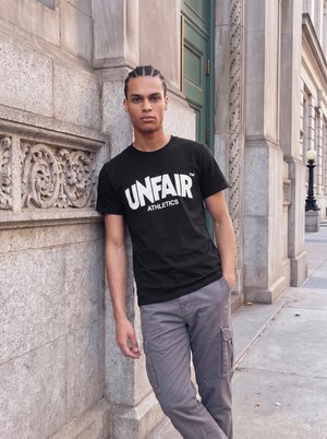Un jeune homme portant un tee-shirt noir UNFAIR Athletics et un pantalon cargo gris est adossé à un bâtiment en pierre sculptée, sur un trottoir en ville.