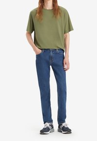 Olivgrön kortärmad t-shirt i kombination med blåjeans. T-shirten har en avslappnad passform; jeansen har en blekt struktur. Grå sneakers med vita detaljer.