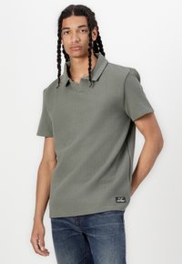 Smaragdgrünes Poloshirt aus strukturiertem Stoff, mit kurzen Ärmeln und einem Kragen. Verfügt über ein kleines schwarzes Etikett auf der Seite.