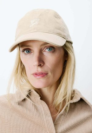 Femme aux yeux bleus portant une casquette de baseball en velours côtelé beige et une chemise assortie, regardant directement la caméra sur un fond uni.