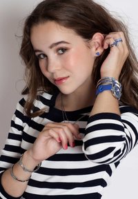 Gestreept marineblauw en witte shirt, zilveren sieraden, blauwe armband en horloge. Oorbellen in de vorm van een hart. Warme huidtinten en natuurlijke make-up.