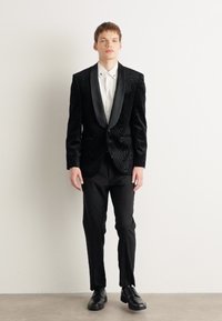 Svart sammetstuxedojacka med texturerat mönster, satinklappor, vit skjorta och svarta byxor. Svarta skor fulländar outfiten.