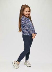 Dziewczyna ubrana w niebieską i fioletową koszulę z długim rękawem w wzory, granatowe legginsy i jasne sneakersy z szarymi akcentami, stoi z złożonymi dłońmi.