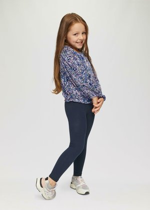 Meisje in een blauw en paars gestreept shirt met lange mouwen, marineblauwe leggings en lichte sneakers met grijze accenten, staande met de handen vouwt.