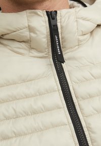 Beige padded jacket featuring a high collar, black zipper with branding, and horizontal quilted texture for insulation.
