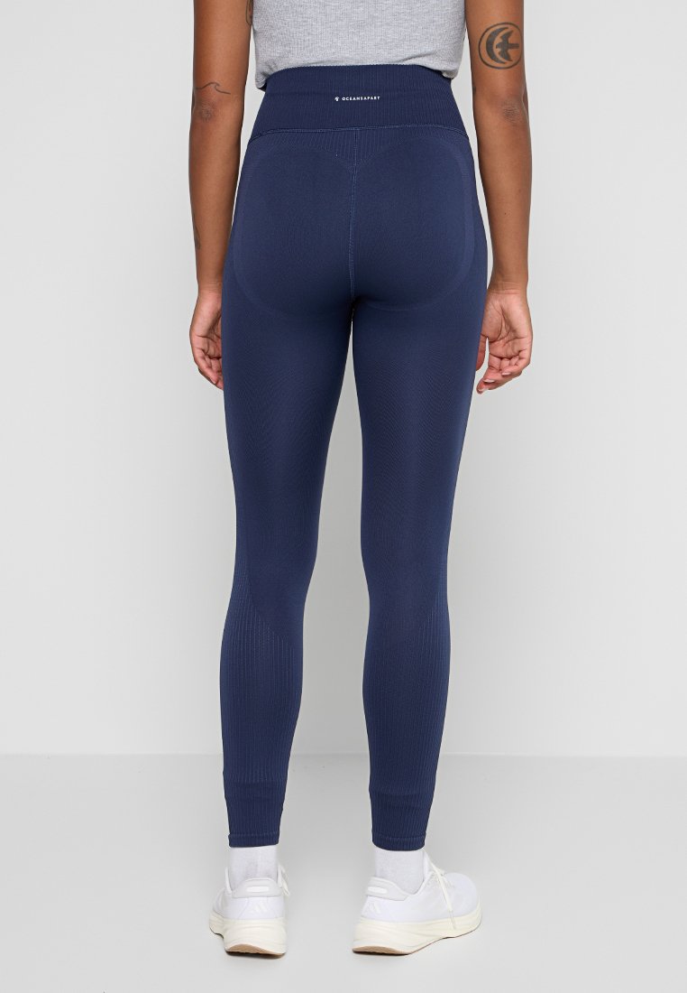 Person, der navyblaue nahtlose Leggings, ein graues ärmelloses Top, weiße Socken und weiße Sneakers trägt, und mit dem Rücken zu einer einfarbigen Hintergrund steht.