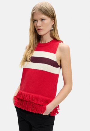 Kobieta o długich blond włosach, ubrana w bezrękawnik czerwony sweter z białymi i ciemnoczerwonymi paskami oraz frędzlami na dole, ręce w kieszeniach czarnych spodni.