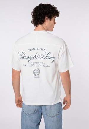 Man with curly hair wearing white oversized T-shirt with "RUNNERS CLUB Classy & Strong" text and light blue jeans, facing away.