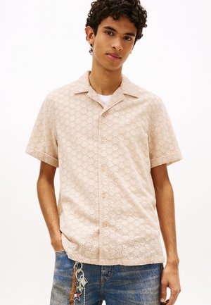 Jonge man draagt een beige kortärmelig overhemd met kantpatroon en blauwe spijkerbroek met sleutelhanger aan de zak, staand met één hand in de zak.