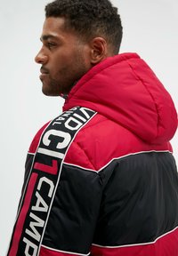 Rote und schwarze Daunenjacke mit Kapuze, die ein auffälliges Logo-Band auf der Schulter und kontrastierende weiße Paspeldetails aufweist.