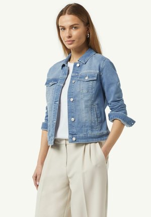 Lichtblauwe spijkerjack met knoopsluiting, twee borstzakken en opgerolde mouwen. Gepaard met een wit T-shirt en beige broek.