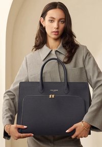 Navy leather tote bag with dual handles, textured finish, front zip pocket, and gold hardware accents. Model holds bag against gray outfit.