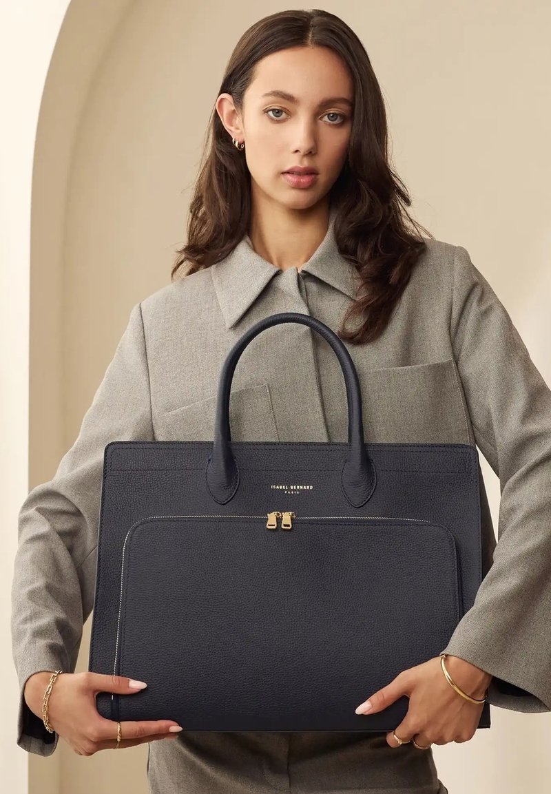 Navy leather tote bag with dual handles, textured finish, front zip pocket, and gold hardware accents. Model holds bag against gray outfit.
