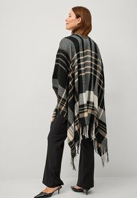 Plaid poncho in black, gray, and beige with fringe at the bottom; made of soft fabric, featuring a loose fit and geometric patterns.