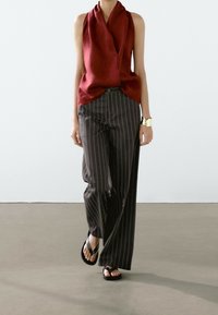 Personne marchant portant un haut rouge sans manches à col bénitier, un pantalon large noir à fines rayures, des sandales noires et un bracelet manchette doré au poignet gauche.