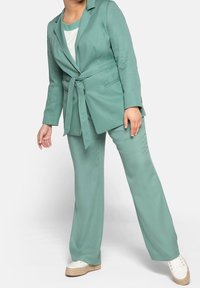 Completo verde menta con giacca con cintura e pantaloni a gamba larga, caratterizzato da una texture liscia e top abbinato. Indossato con scarpe bianche.