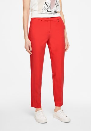 Femme portant un pantalon rouge vif ajusté, des baskets blanches et un haut sans manches blanc avec un motif noir et du texte.