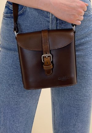 Person, der eine dunkelbraune Leder-Crossbody-Tasche mit Schnallenverschluss hält und helle blaue Denim-Jeans trägt.