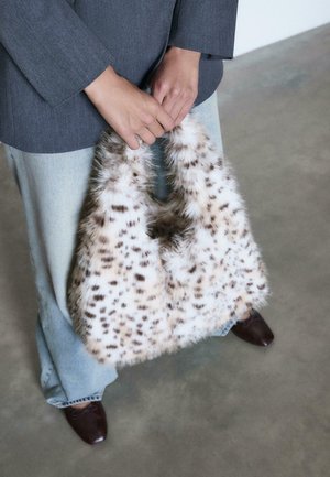 Hands holding a white and brown spotted faux fur bag, wearing a gray blazer, light blue jeans, and dark brown shoes on a concrete floor.
