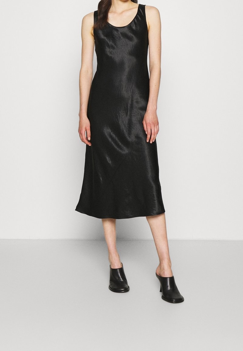 Femme portant une robe midi en satin noire sans manches et des mules noires à talons, debout devant un fond blanc uni.