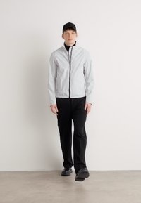 Ανοιχτό γκρι ζακέτα με φερμουάρ και όρθιο γιακά, μαύρο φερμουάρ και λογότυπα, σε συνδυασμό με μαύρο παντελόνι και μαύρο καπέλο, καθισμένος μπροστά σε έναν λευκό τοίχο.