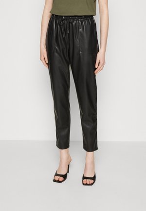 Woman wearing black faux leather tapered pants with elastic waist and black open-toe heeled sandals, standing against a plain background.
