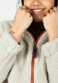 Fuzzy cream hoodie with a zip closure and contrasting red trim. Features prominent texture and a black zipper pull. Includes visible tattoos.