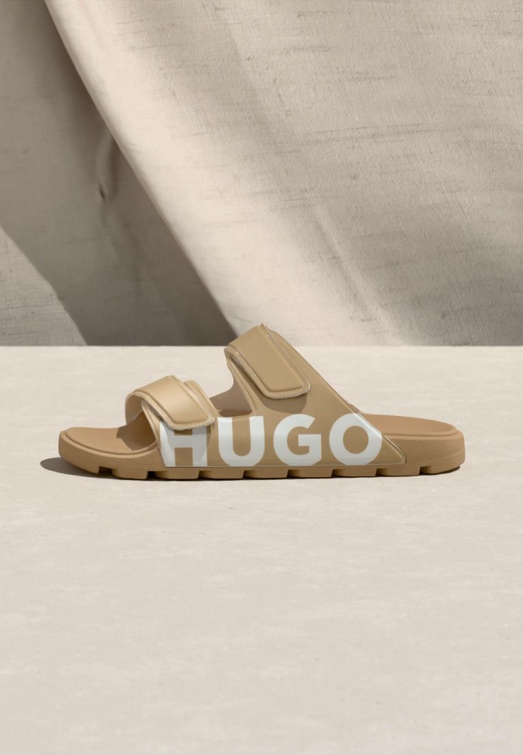 Sandale beige à bout ouvert avec deux lanières réglables et semelle texturée, marquée de grandes lettres blanches "HUGO" sur le côté.