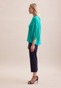 Blusa turchese con maniche a palloncino, abbinata a pantaloni cropped blu navy e sandali neri con tacco alto. Tessuto liscio e linee pulite.