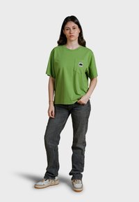 Camiseta de algodón verde con cuello redondo y mangas cortas, que presenta un pequeño bolsillo con un logo de elefante en blanco y negro. Ajuste casual.