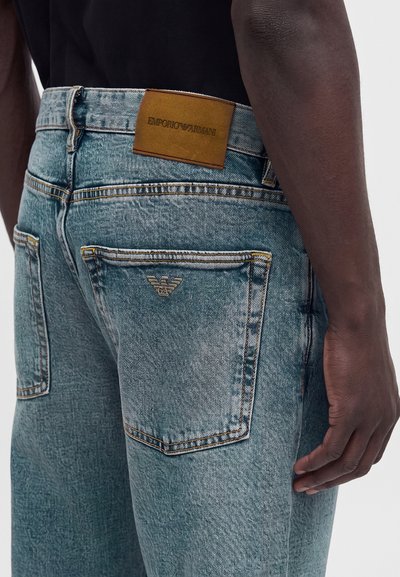 Άνδρας που φορά ανοιχτό μπλε τζιν Emporio Armani με λογότυπο στο πίσω τσέπη και καφέ δερμάτινη ετικέτα στη μέση, εμφανιζόμενος από τη μέση μέχρι το μέσο του μηρού.