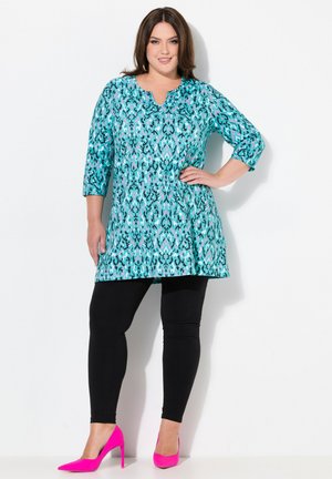 Tunique turquoise avec motif abstrait noir et rose, décolleté en V, manches trois-quarts, associée à un legging noir et des escarpins roses.