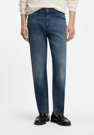 Mann trägt blaue Jeans mit geradem Bein, schwarze Loafers und einen cremefarbenen Pullover, steht vor weißem Hintergrund.