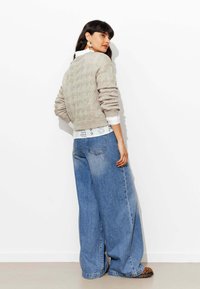 Pull en maille beige, superposé sur un chemisier en dentelle blanche, associé à un jean bleu à jambes larges et des chaussures à imprimé animal. Modèle de dos.