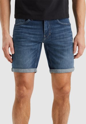 Man in opgerolde blauwe denimshorts met voorzakken en een zwart shirt, staande tegen een effen lichte achtergrond.