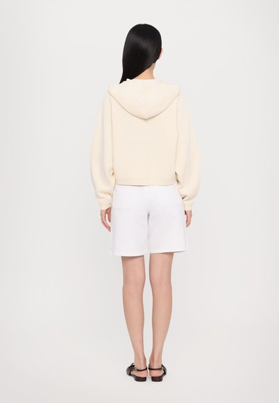 Krémový, skrátený Mikina s kapucňou a textúrovaným vzorom, v kombinácii s bielymi šortkami a čiernymi sandálmi, z pohľadu zozadu.